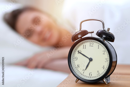 Alarm clock opposite of beautiful young woman sleeping while lying in bed comfortably and blissfully. Happy waking up early for a job