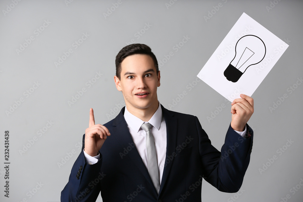 Emotional young businessman holding paper sheet with drawn light bulb as symbol of idea on grey background Emotional young businessman holding paper sheet with drawn light bulb as symbol of idea on grey background