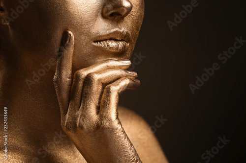 Beautiful young woman with golden paint on her body against dark background, closeup
