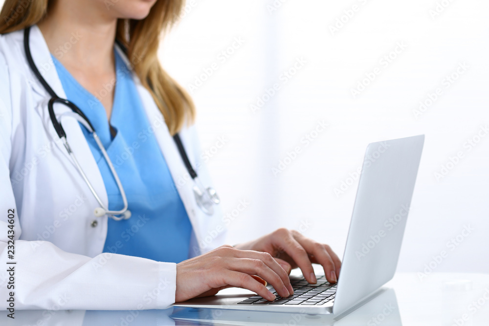 Doctor typing on laptop computer while sitting at the glass desk in hospital office. Physician at work. Medicine and healthcare concept Doctor typing on laptop computer while sitting at the glass desk in hospital office. Physician at work. Medicine and healthcare concept