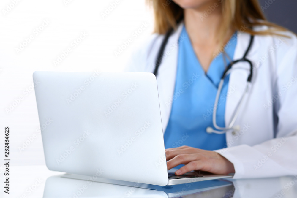 Doctor typing on laptop computer while sitting at the glass desk in hospital office. Physician at work. Medicine and healthcare concept Doctor typing on laptop computer while sitting at the glass desk in hospital office. Physician at work. Medicine and healthcare concept