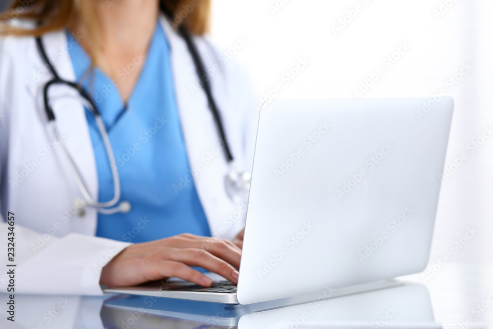 Doctor typing on laptop computer while sitting at the glass desk in hospital office. Physician at work. Medicine and healthcare concept Doctor typing on laptop computer while sitting at the glass desk in hospital office. Physician at work. Medicine and healthcare concept