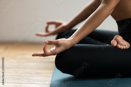 Young sporty yogi woman practicing yoga, doing Ardha Padmasana exercise, Half Lotus pose with mudra gesture, working out, wearing sportswear pants, indoor close up, yoga studio. Well-being concept