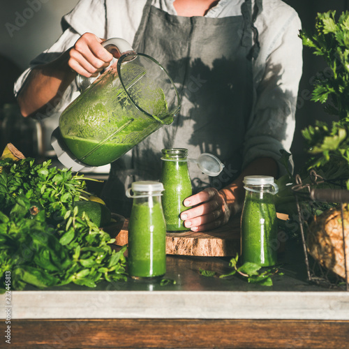 Making green detox take-away smoothie. Woman in linen apron pouring green smoothie drink from blender to bottle surrounded with vegetables and greens, square crop. Healthy, weight loss food concept