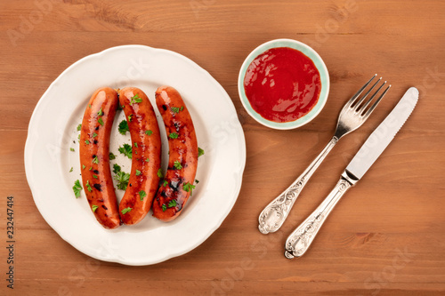 A photo of a plate of fried sausages with ketchup, shot from the top on a rustic wooden background with a fork and a knife, with a place for text