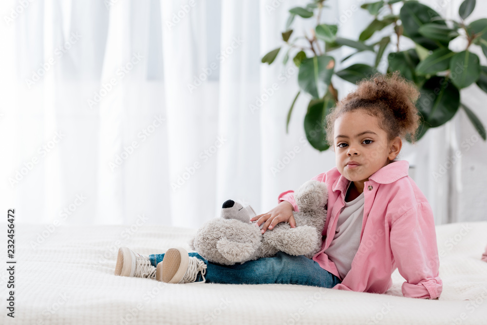 Upset african american kid sitting on the bed with teddy bear on her knees Upset african american kid sitting on the bed with teddy bear on her knees