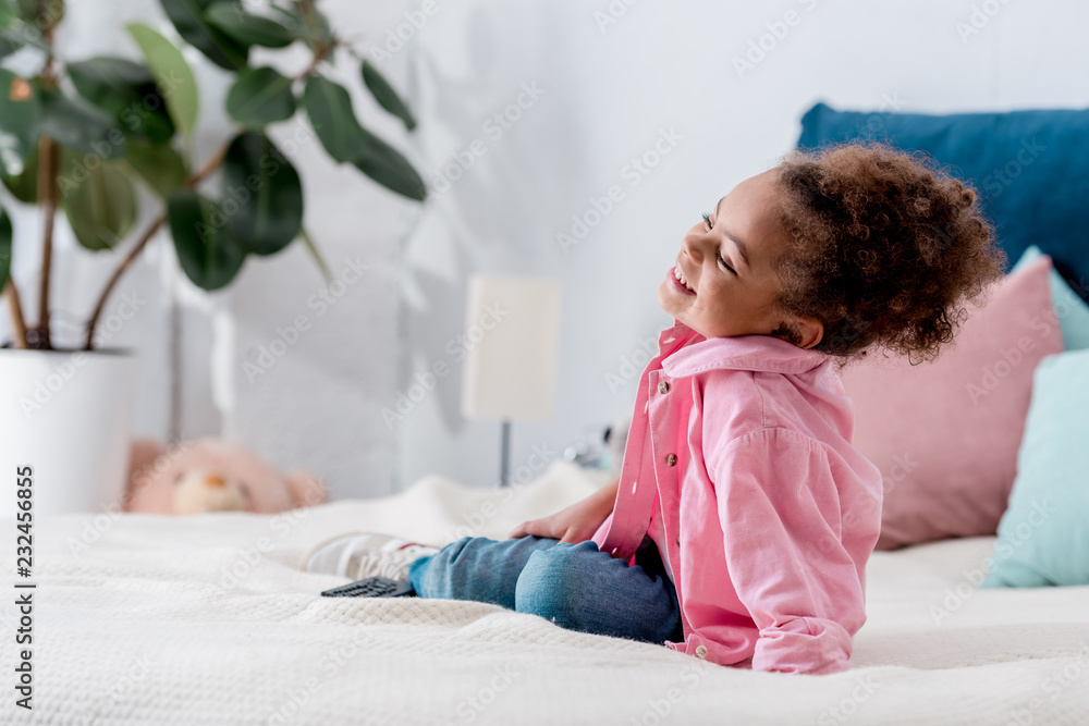Fototapeta premium Curly African american kid sitting on the bed and smiling