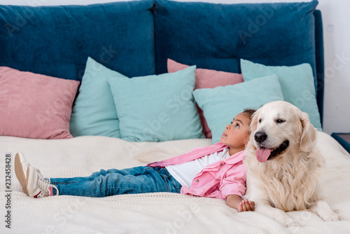 Adorable little african american kid in pink jacket lying on bed and leaning to dog