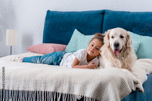 Cute little african american kid with happy dog lying on bed with colourful pillows photo