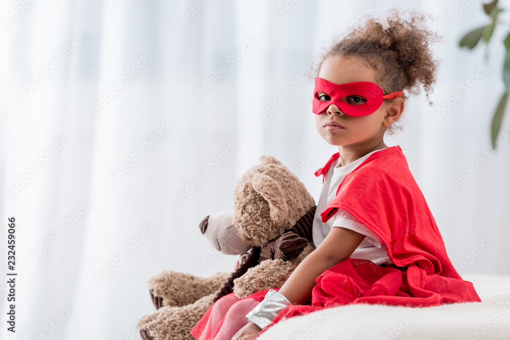 Cute african american child in red superhero costume and mask with teddy bear looking at camera Cute african american child in red superhero costume and mask with teddy bear looking at camera