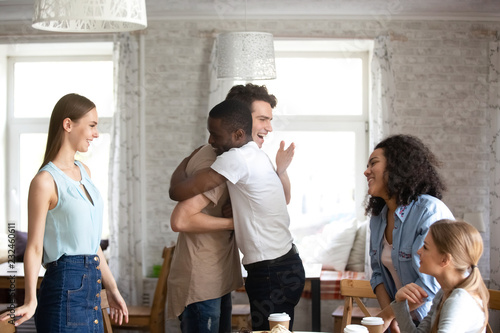 Diverse students gathered together during lunch. Guys buddies best friends hugging greeting each other at meeting in cafe surrounded by attractive multi-ethnic girls. Friendship and reunion concept