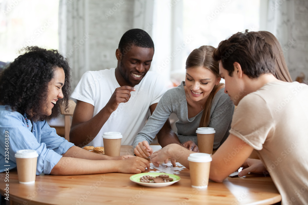 Diverse multiracial friends cheerful girls and guys drinking coffee having fun laughing assembling puzzle jigsaw sitting together at table in home or cafeteria. Teamwork and weekend activities concept Diverse multiracial friends cheerful girls and guys drinking coffee having fun laughing assembling puzzle jigsaw sitting together at table in home or cafeteria. Teamwork and weekend activities concept