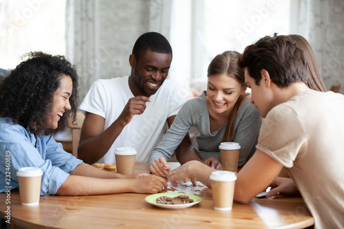 Diverse multiracial friends cheerful girls and guys drinking coffee having fun laughing assembling puzzle jigsaw sitting together at table in home or cafeteria. Teamwork and weekend activities concept
