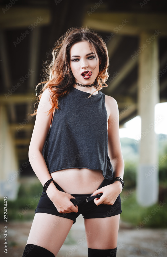 Outdoor shot: sexy young rock girl in black shorts and shirt standing under  bridge and showing tongue Stock Photo | Adobe Stock