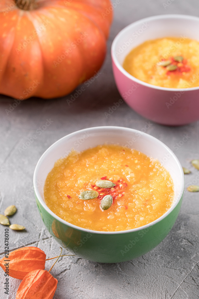 Pumpkin soup puree in ceramic bowls and orange large pumpkin on a gray textured background. Pumpkin soup puree in ceramic bowls and orange large pumpkin on a gray textured background.