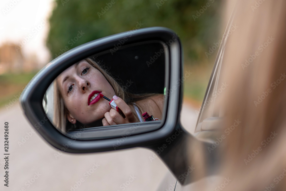 A young, beautiful woman with long hair looks in the car's rearview mirror and paints her lips.