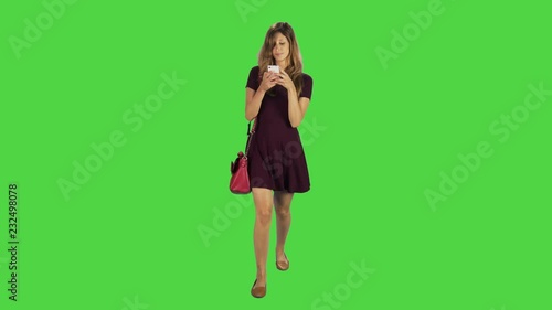 A young woman walking towards camera, texting gladly in a full body shot over a green screen.