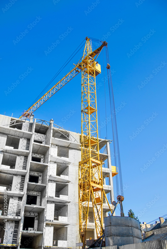 Yellow construction crane at a construction site near an unfinished building of concrete blocks on a background of blue sky. Construction technologies, expansion of cities, employment, job search