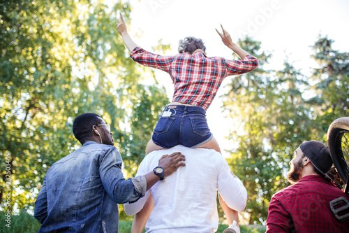 Back side of unrecognizable mixed race diverse young people going at the park in summer afternoon. Boyfriend carrying his girlfriend on piggyback and one of guys carries bmx byke over his shoulder. photo