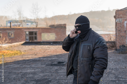 a man, a terrorist, a bandit in a black leather jacket and a mask talking on the phone on the street near an abandoned building