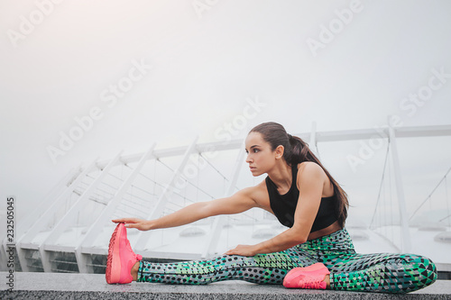 Picture of well-built model stratching outside. She sits and leans forward. Young woman is serious and concentrated. She does exercise at big white stadium.
