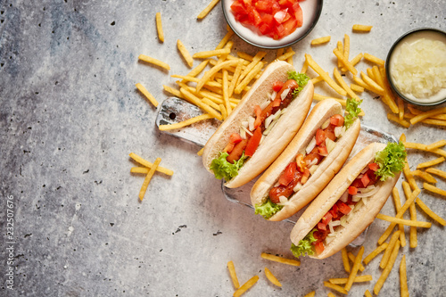 Assortment of three tasety hot dogs, placed on wooden cutting board. Served with french fries, onion and tomato. View from above. Stone background. photo