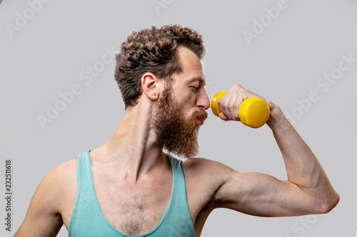 Bodybuilding and Sports concept. Skinny adult bearded man in singlet, slim mature man with anorexic body comically demonstrates his musculs with raised hands and dumbbell photo