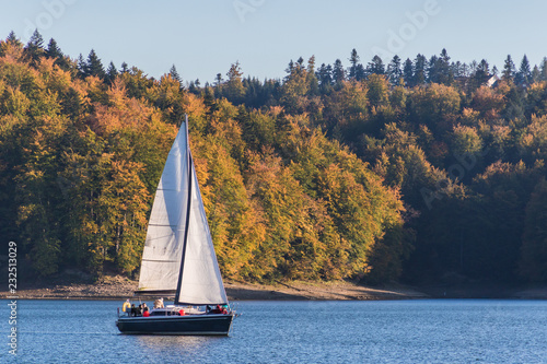 Autumnal landscape with one sailboat sailing on the lake surrounded by hill grown with forest trees on a sunny day