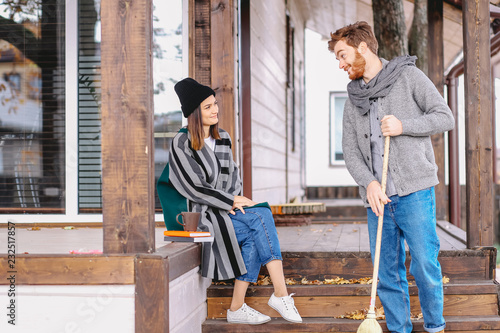 A handsome european male dressed in warm knitted sweater and scarf cleaning a wooden porch with a broom, sweeping autumn leaves, while his wife entertaing him with funny stories photo