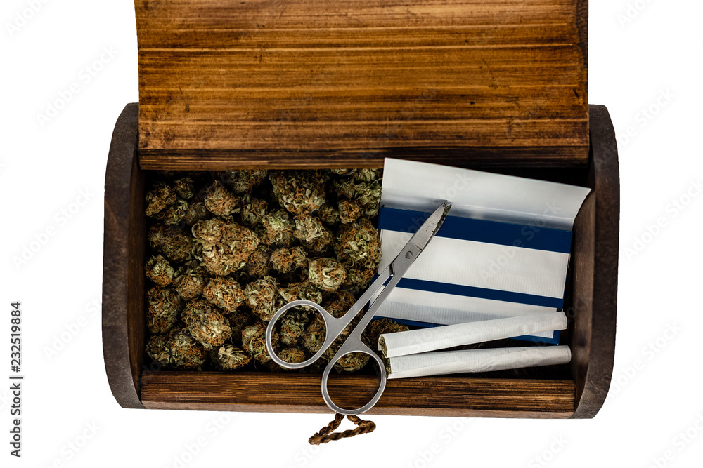 A wooden stash box half filled with green marijuana buds covered in rich red hairs with a pair of scissors, 2 prerolled joints & a pack of rolling papers on a white background.