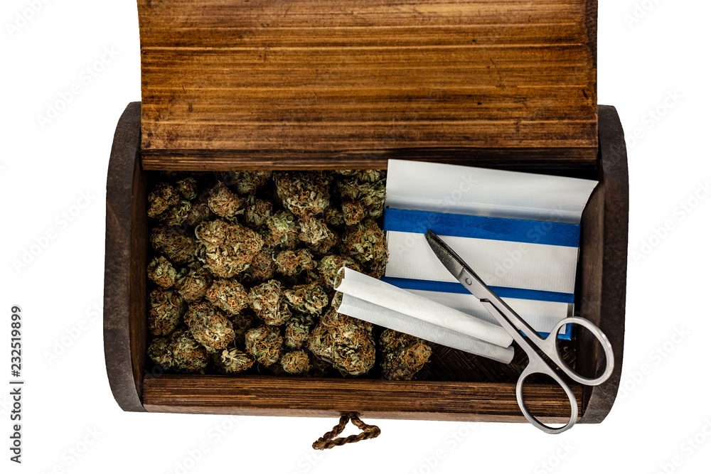 A wooden stash box half filled with green marijuana buds covered in rich red hairs with a pair of scissors, 2 prerolled joints & a pack of rolling papers on a white background.