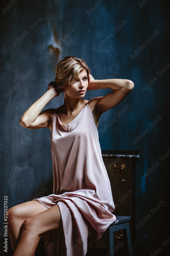 Portrait of sensual blonde woman in a long silk evening dress, fashion beauty photo in a simple interior