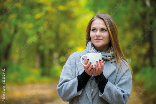 beautiful girl in a gray coat holding a mug in her palms. walk in the autumn park. Woman happy with cup of coffee in hands. Happy woman drinking cofee to go outdoors in autumn park