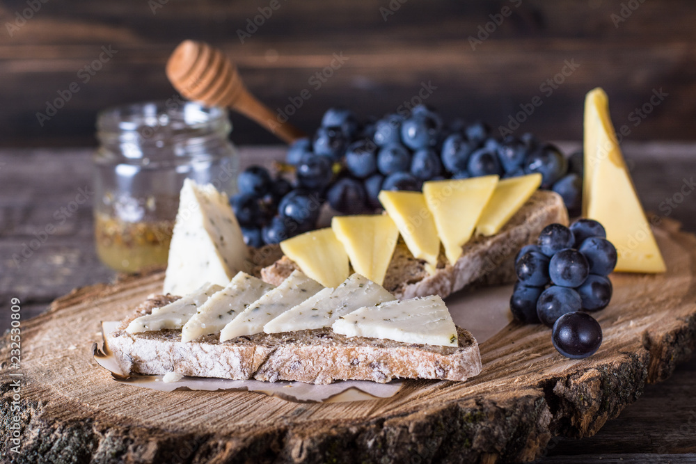 Assorted cheeses with grapes, bread, honey on dark wood background. Goat cheese with herbs. Natural wooden board. Italian appetizer. bruschetta with cheese. Still life of food. Breakfast concept.