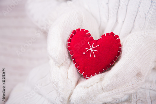 Hands in white knitted mittens holding red decorative heart.