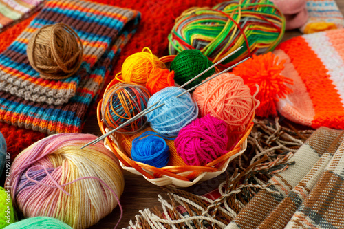 Colorful yarn for knitting clothes. Multicolored yarn for knitting in a basket surrounded by a plaid, a hat and a scarf. Knitting is a type of needlework.