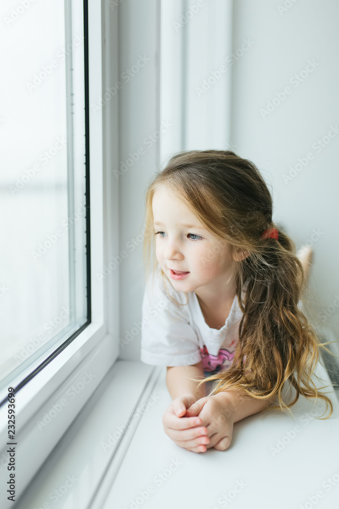 Beautiful little girl smiling and watching out the window. A child looks out the window. Young girl looking from window. Portrait of cheerful kid sits at window. Beautiful little girl smiling and watching out the window. A child looks out the window. Young girl looking from window. Portrait of cheerful kid sits at window.