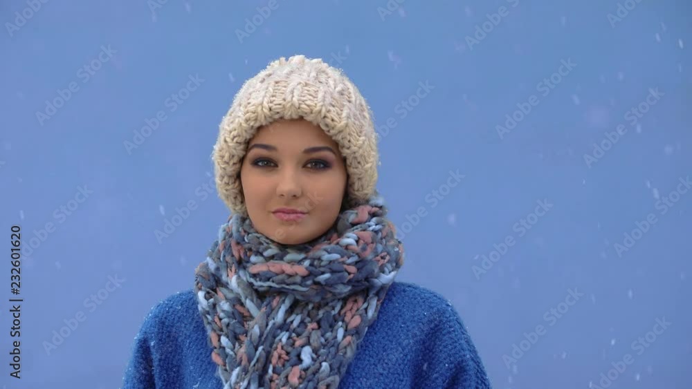 Serious women facing camera during winter holidays outside. Snow falling in slow motion 4K. Young confident female enjoying winter day outdoors. Woman in cap, coat, scarf standing against blue copy Serious women facing camera during winter holidays outside. Snow falling in slow motion 4K. Young confident female enjoying winter day outdoors. Woman in cap, coat, scarf standing against blue copy