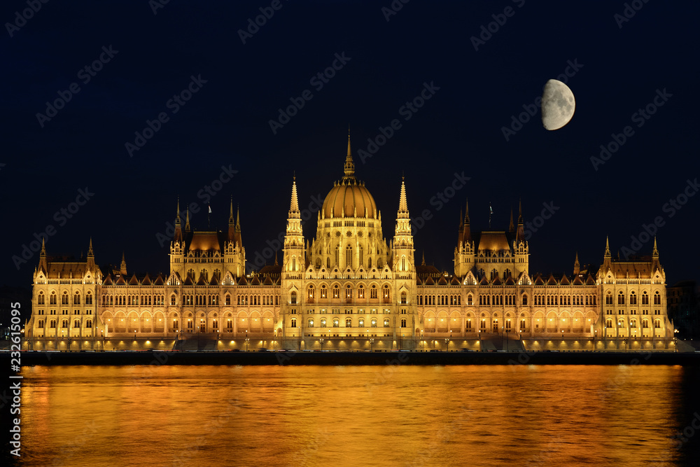 Night cityscape of illuminated Budapest parliament building with moon and golden reflection in Danube river