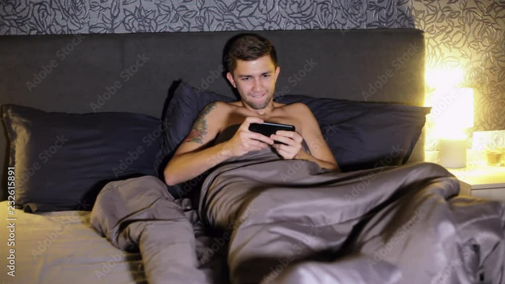 Young man playing games on smartphone while lying in bed at home in the evening. Tensely plays, wins the game and is very happy Young man playing games on smartphone while lying in bed at home in the evening. Tensely plays, wins the game and is very happy