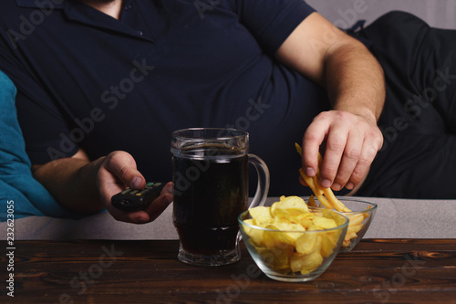overeating, sedentary lifestyle, bad habits, food addiction, eating disorders. fat overweight man lay on the coach with tv remote, junk food and beer. depression, laziness alcohol addiction