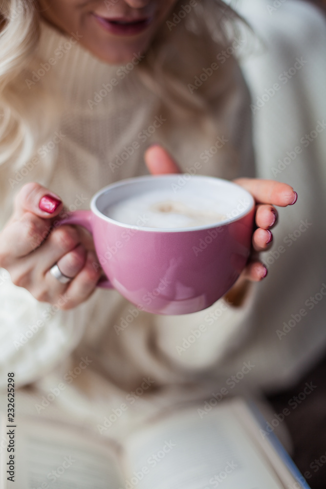 Cozy home portrait of a young beautiful woman. A beautiful blonde girl in a knitted dress is sitting by the window drinking a cappuccino and reading a book. Autumn decor. Cozy. Winter. Cozy home portrait of a young beautiful woman. A beautiful blonde girl in a knitted dress is sitting by the window drinking a cappuccino and reading a book. Autumn decor. Cozy. Winter.