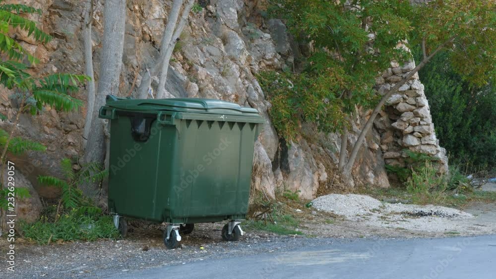 A dark green plastic trash bin on wheels stands next to the road in the mountains. A dark green plastic trash bin on wheels stands next to the road in the mountains.