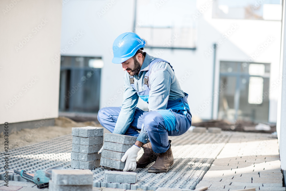 Builder in uniform laying paving tiles on the construction site with white houses on the background