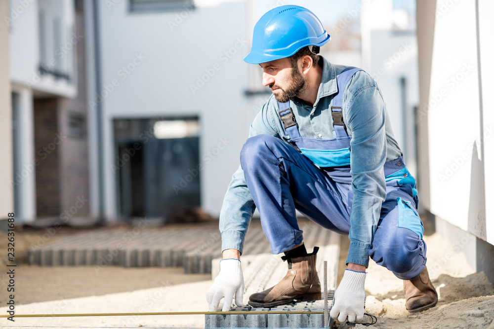 Builder in uniform mounting paving tiles on the construction site with white houses on the background