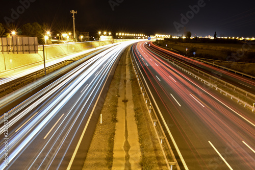 DIFFERENT WHITE, BLUE AND RED LIGHTS OF MANY VEHICLES DRIVING IN A CURVE OF A SPANISH MOTORWAY WITH THE LIGHT OF A CITY AT THE END OF THE HORIZON