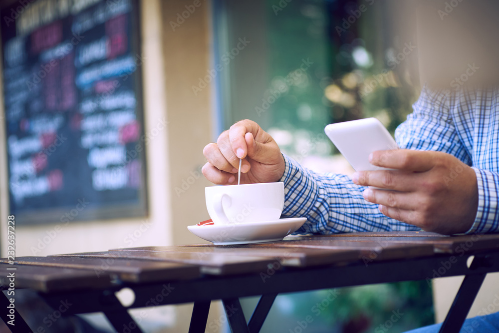 Always in touch with his colleagues. Close-up of man holding mobile phone and coffee cup while sitting on the terrace of the summer cafe.