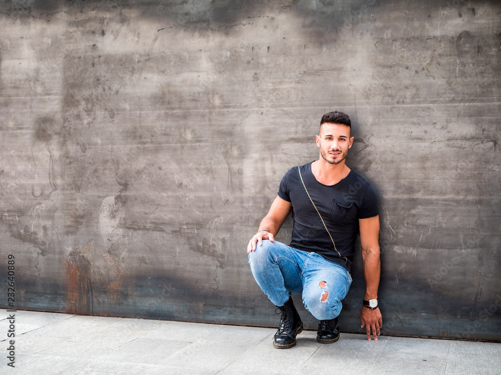 Handsome young man standing against concrete wall, looking at camera, wearing black t-shirt and jeans. Full length shot