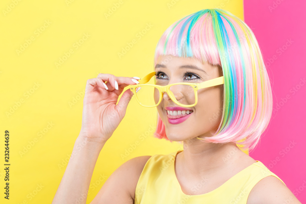 Young woman in a colorful wig with sunglasses on a split yellow and pink background Young woman in a colorful wig with sunglasses on a split yellow and pink background