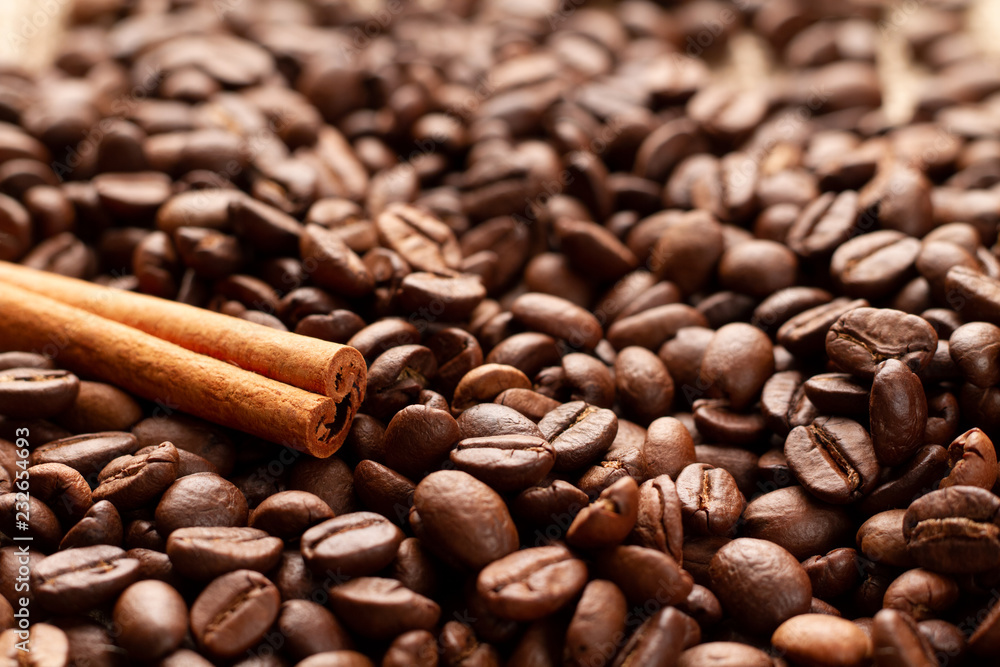 Fototapeta premium A gorgeous stick of natural fragrant brown cinnamon lies on freshly roasted coffee beans in the lower left third of the frame in a golden section. Photo of coffee beans and cinnamon in backlight.